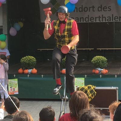 Das Bild zeigt einen jonglierenden Clown auf einem Einrad vor einer Bühne. Im Vordergrund sind mehrere Kinder zu sehen.
