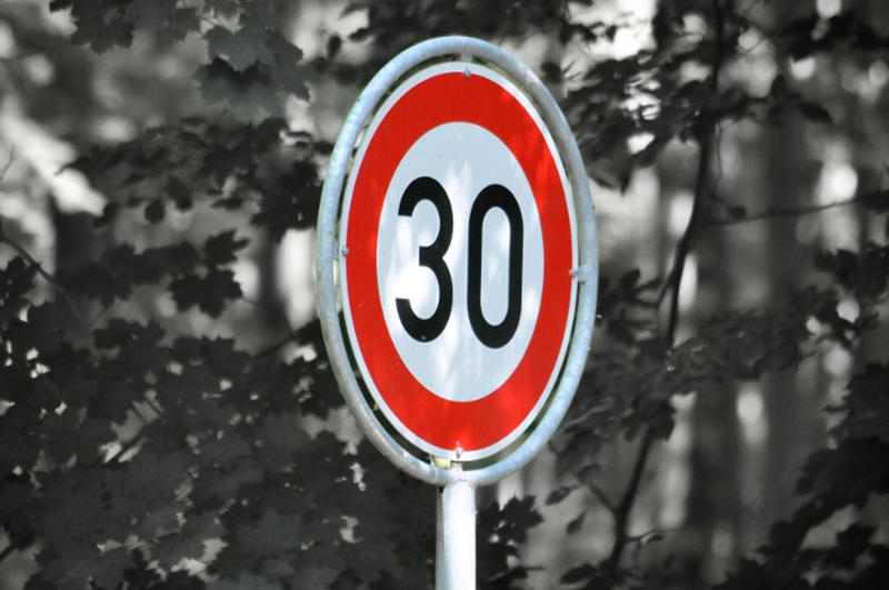 Vor schwarz-weißem Hintergrund ist ein rot-weißes Tempo 30 Schild zu erkennen.