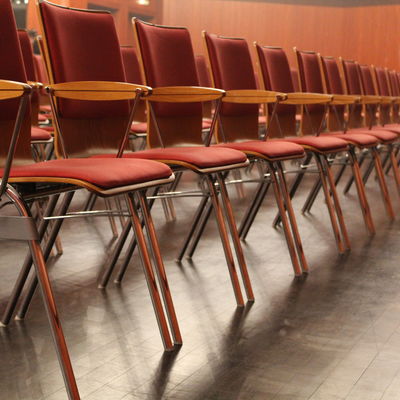Erste Reihe der Reihenbestuhlung in der Stadthalle. Metalrahmen mit hellem Holz und roten Sitzpolstern.