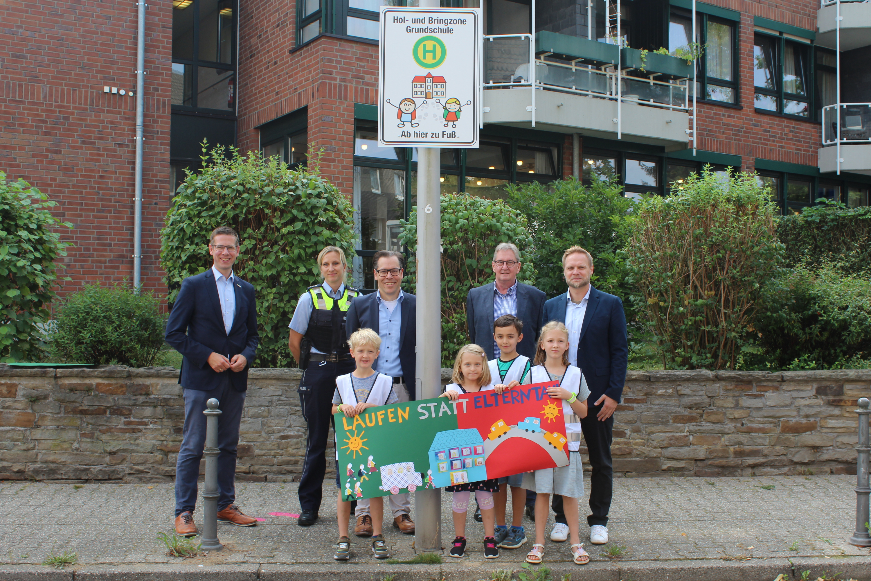Bürgermeister Christoph Schultz gemeinsam mit Erwachsenen und Kindern an einem Schild mit der Aufschrift "Hol- und Bringzone Grundschule"