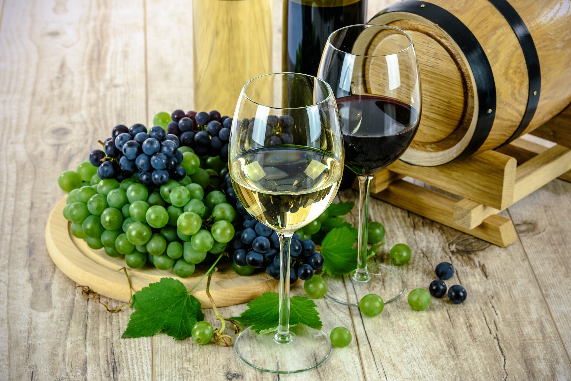 Das Bild zeigt eine Zusammenstellung mehrerer Gegenstände, die mit Wein zu tun haben. Einige Reben in grün und rot, zwei gefüllte Weingläser und ein kleines Tischfass.