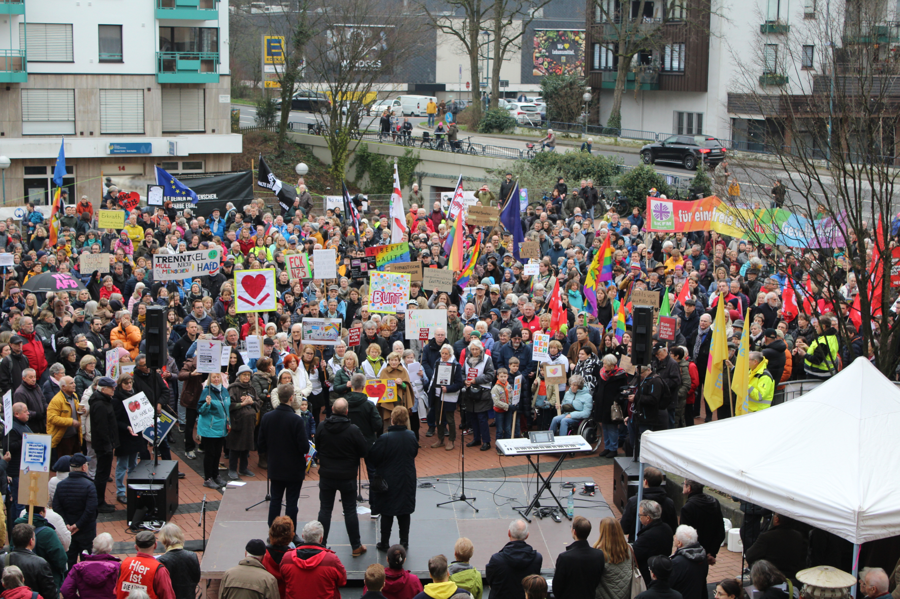 Demonstration mit vielen Menschen mit bunten Fahnen und Plakaten stehen vor einer kleinen Bühne, wo jemand spricht.
