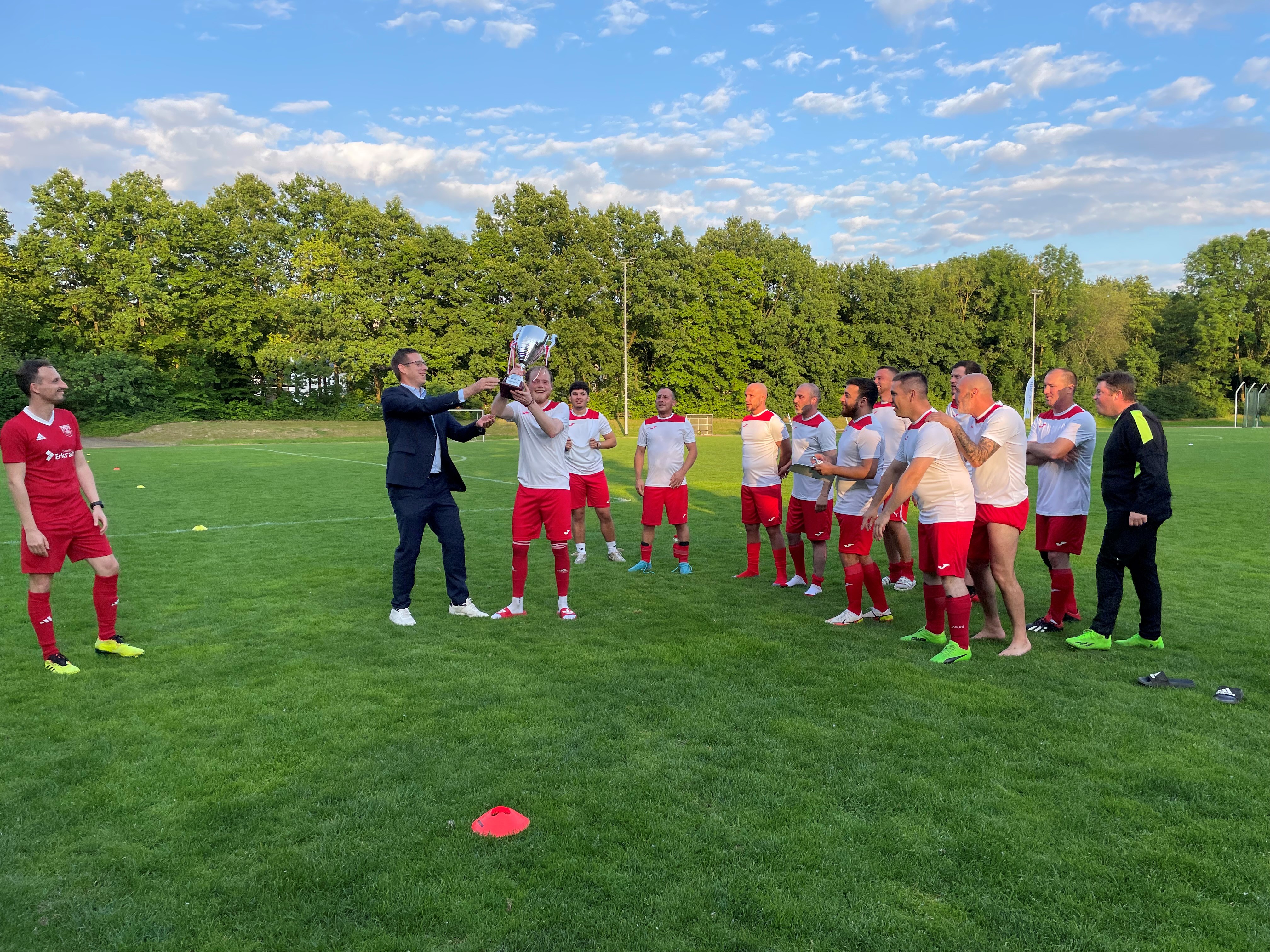 Das Bild zeigt eine Fußballmannschaft auf einem Rasenplatz, die einen Sieg feiert. Der Bürgermeister überreicht dem Team einen Pokal.
