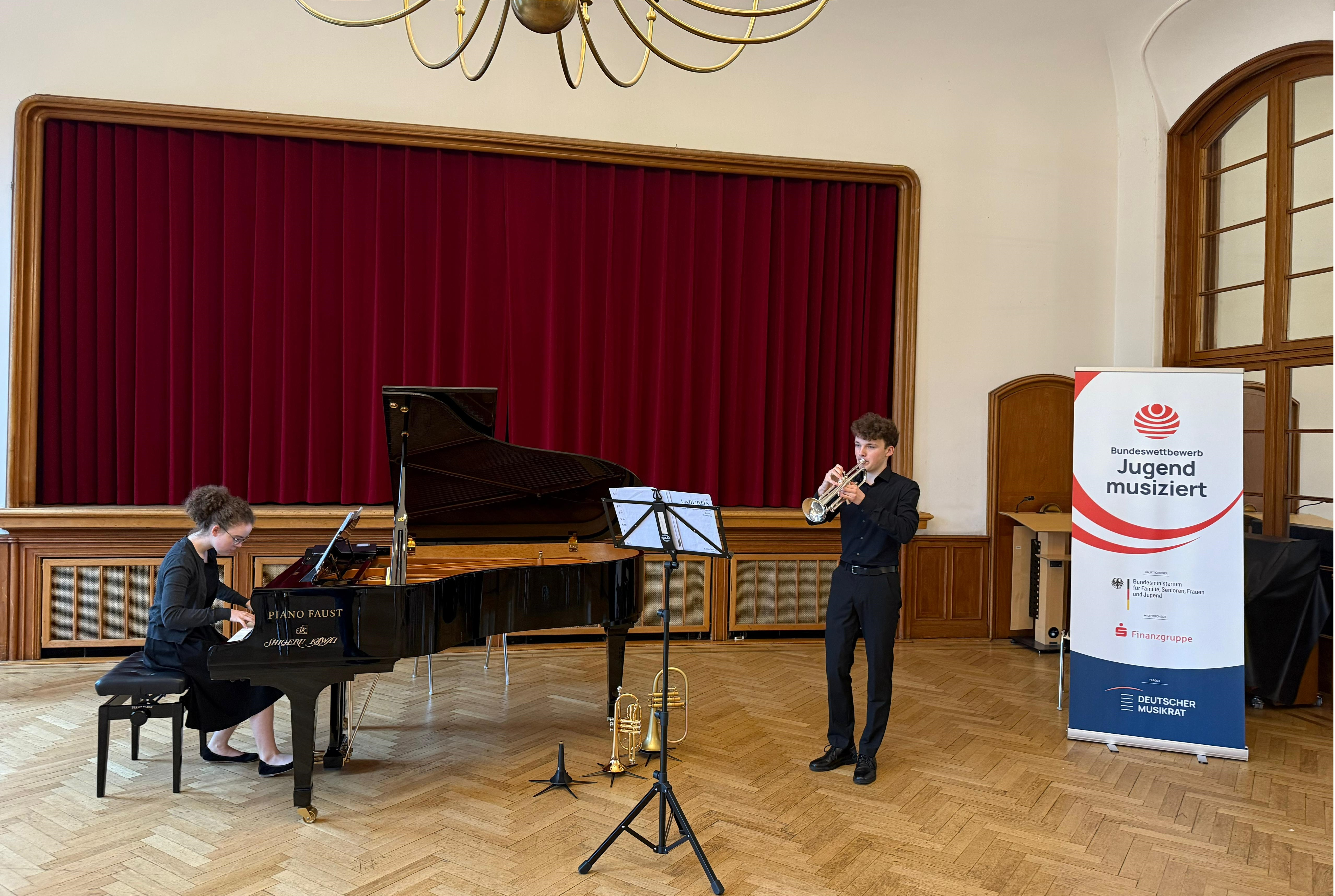 JMS Erkrath erfolgreich bei Jugend musiziert Eine junge Frau im schwarzen Kleid spielt in einem Saal vor einer Bühne an einem Flügel, daneben spielt ein junger Mann in schwarzem Hemd und Hose Trompete. Neben ihnen am rechten Bildrand steht ein Banner des Wettbewerbs "Jugend musiziert".