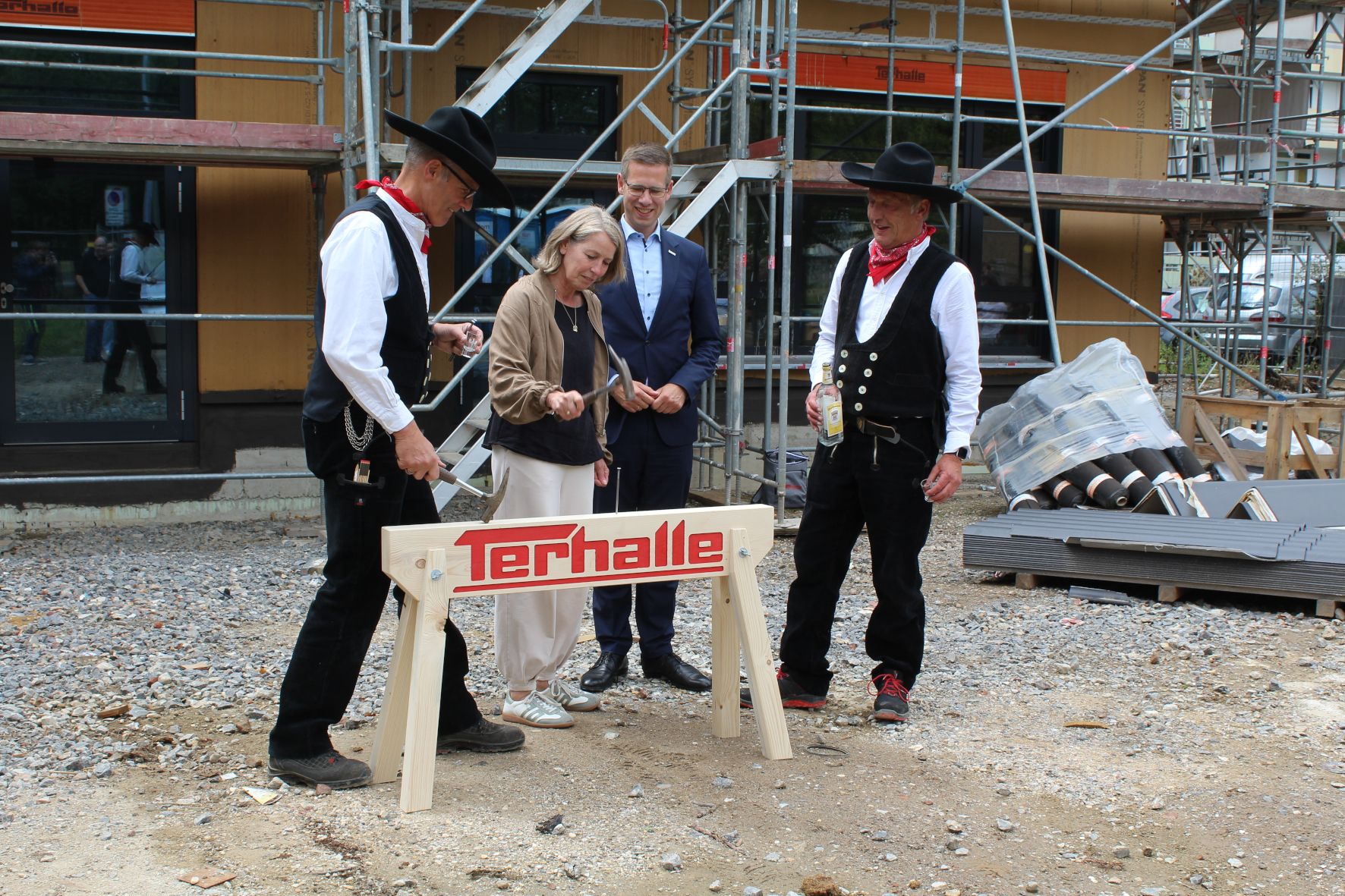 Zwei Zimmerleute in Tracht stehen mit einer Frau und einem Mann vor einem Holzbalken. Die Frau schlägt zaghaft einen Nagel mit einem Zimmermannshammer ein, der Mann dahinter lächelt.