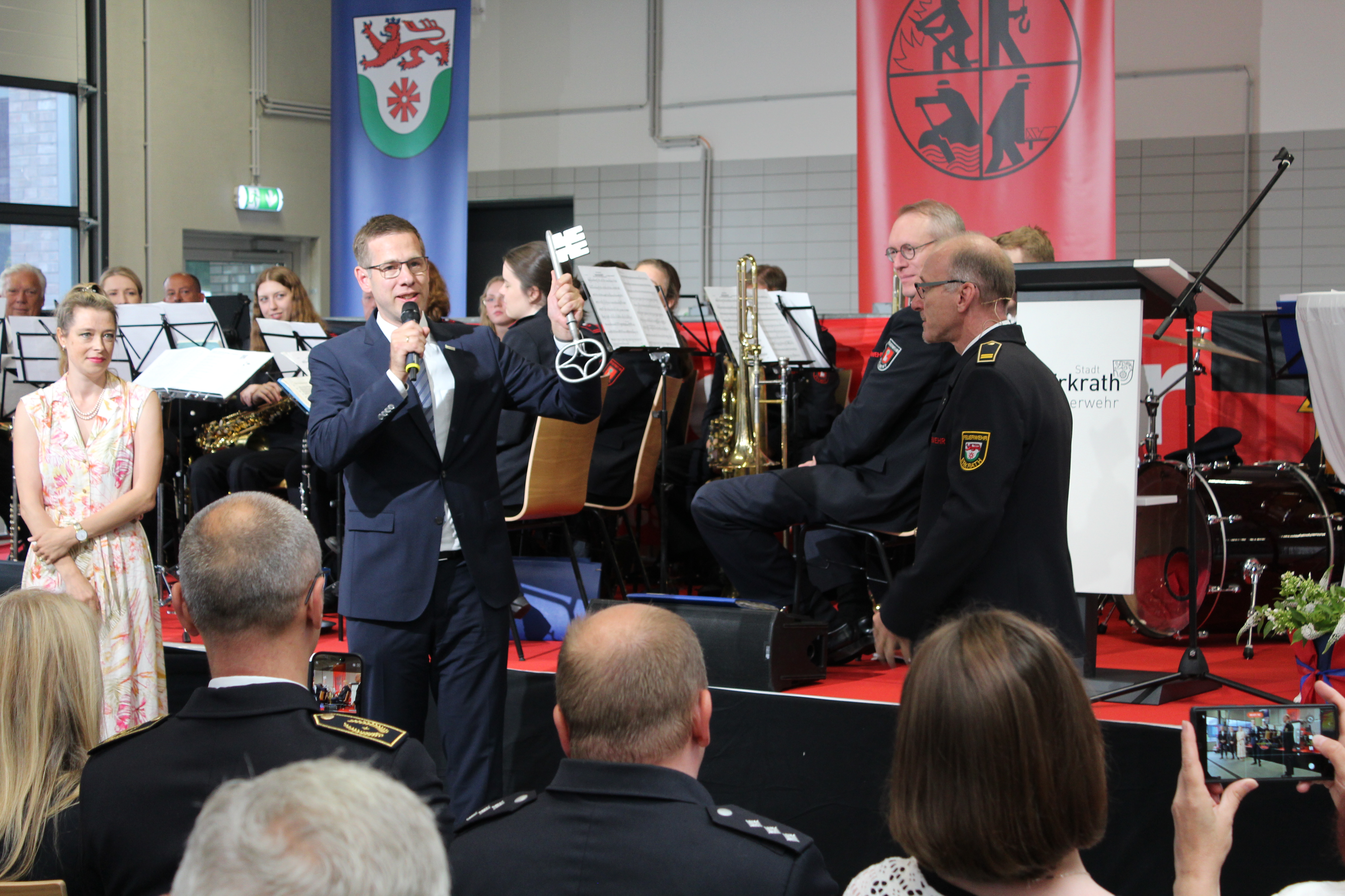Bürgermeister Christoph Schultz (m.) übergibt einen großen Schlüssel an Feuerwehrchef Guido Vogt (r.). Im Hintergrund sitzt ein Orchester auf einer Bühne. Im Vordergrund ist das Publikum im Anschnitt zu sehen.