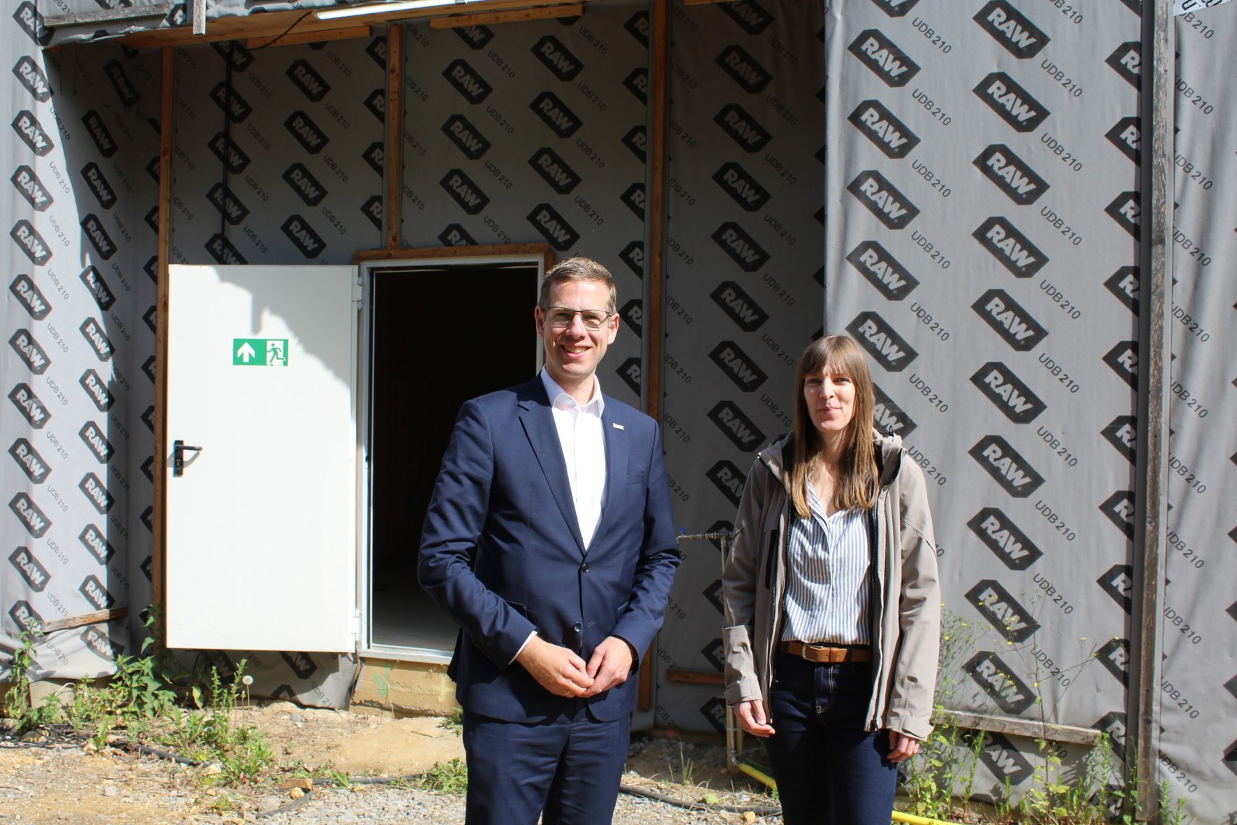 Bürgermeister Schultz und eine Mitarbeiterin stehen vor dem verkleideten Rohbau der Kita Karlstraße