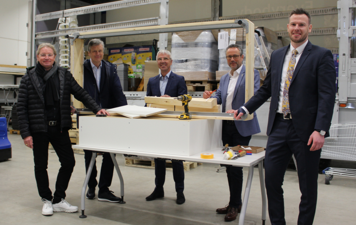 Fünf Personen stehen in einer Halle. Vor ihnen befindet sich Werkzeug und ein Möbelstück zum aufbauen.