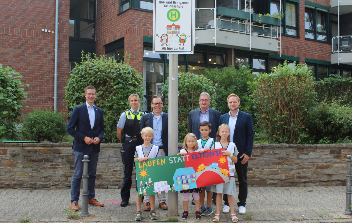 Bürgermeister Christoph Schultz gemeinsam mit Erwachsenen und Kindern an einem Schild mit der Aufschrift "Hol- und Bringzone Grundschule"