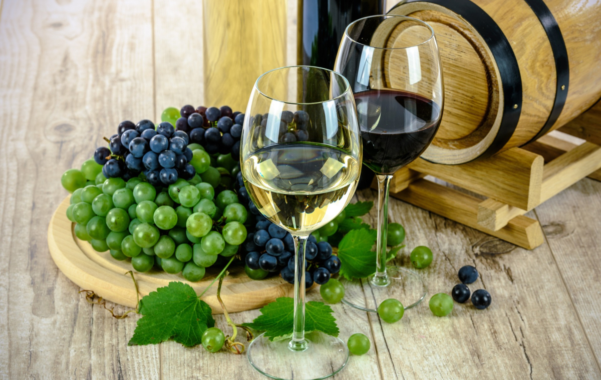 Das Bild zeigt eine Zusammenstellung mehrerer Gegenstände, die mit Wein zu tun haben. Einige Reben in grün und rot, zwei gefüllte Weingläser und ein kleines Tischfass.