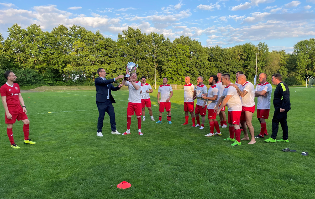 Das Bild zeigt eine Fußballmannschaft auf einem Rasenplatz, die einen Sieg feiert. Der Bürgermeister überreicht dem Team einen Pokal.