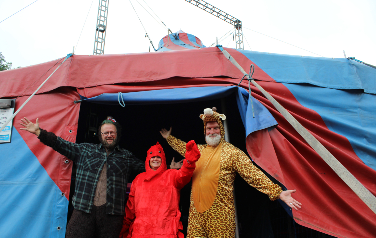 Drei Personen stehen im Eingang eines rot-blauen Zirkuszelt. Einer ist als Clown, einer als Hummer und einer als Leopard verkleidet