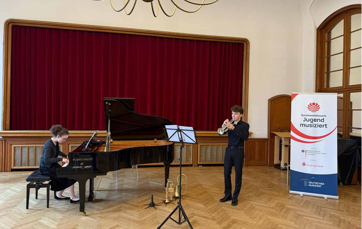 Eine junge Frau im schwarzen Kleid spielt in einem Saal vor einer Bühne an einem Flügel, daneben spielt ein junger Mann in schwarzem Hemd und Hose Trompete. Neben ihnen am rechten Bildrand steht ein Banner des Wettbewerbs "Jugend musiziert".