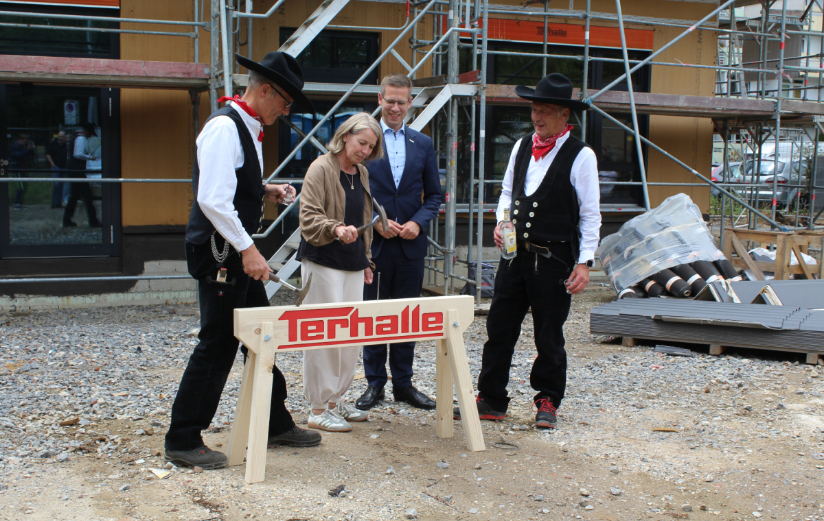 Zwei Zimmerleute in Tracht stehen mit einer Frau und einem Mann vor einem Holzbalken. Die Frau schlägt zaghaft einen Nagel mit einem Zimmermannshammer ein, der Mann dahinter lächelt.