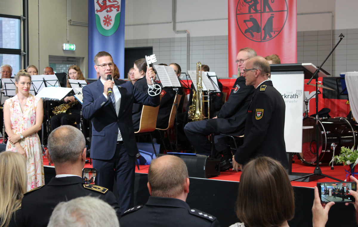 Bürgermeister Christoph Schultz (m.) übergibt einen großen Schlüssel an Feuerwehrchef Guido Vogt (r.). Im Hintergrund sitzt ein Orchester auf einer Bühne. Im Vordergrund ist das Publikum im Anschnitt zu sehen.
