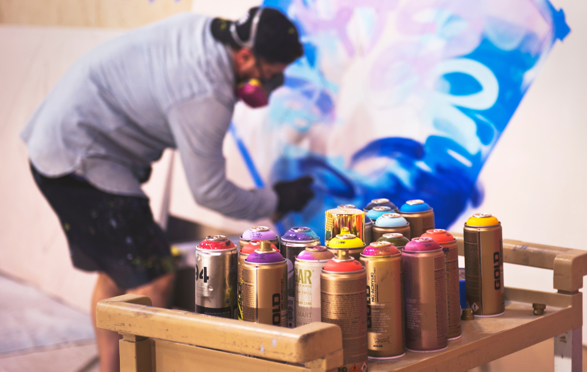Graffiti Spraydosen im Vordergrund. Im Hintergrund eine Person, die ein Graffiti auf eine Leinwand sprüht.