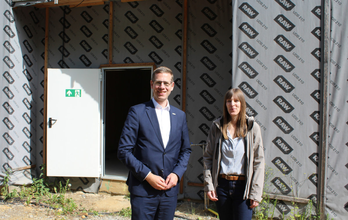 Bürgermeister Schultz und eine Mitarbeiterin stehen vor dem verkleideten Rohbau der Kita Karlstraße